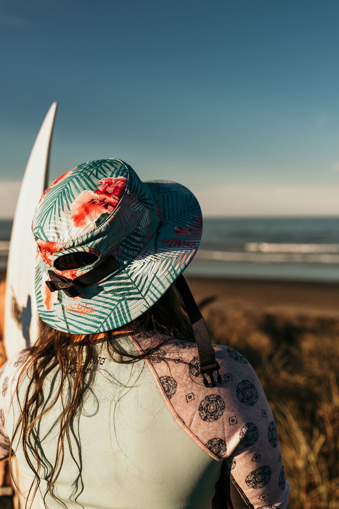 Tropical print bucket hat resting on a surfboard with ocean views in the background.