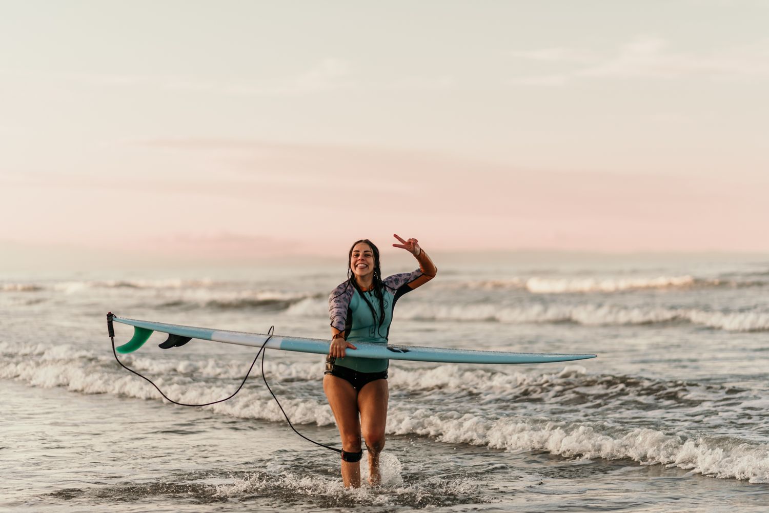 Surfer walking through shallow waves while holding a surfboard and waving.