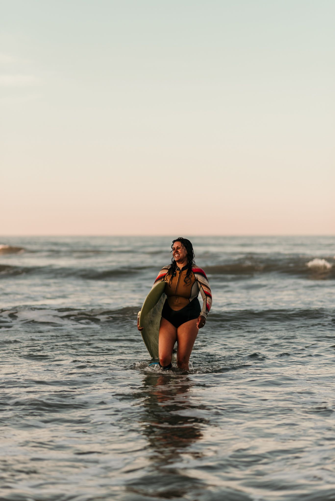 Surfer wading through ocean waves while carrying a surfboard at sunset.