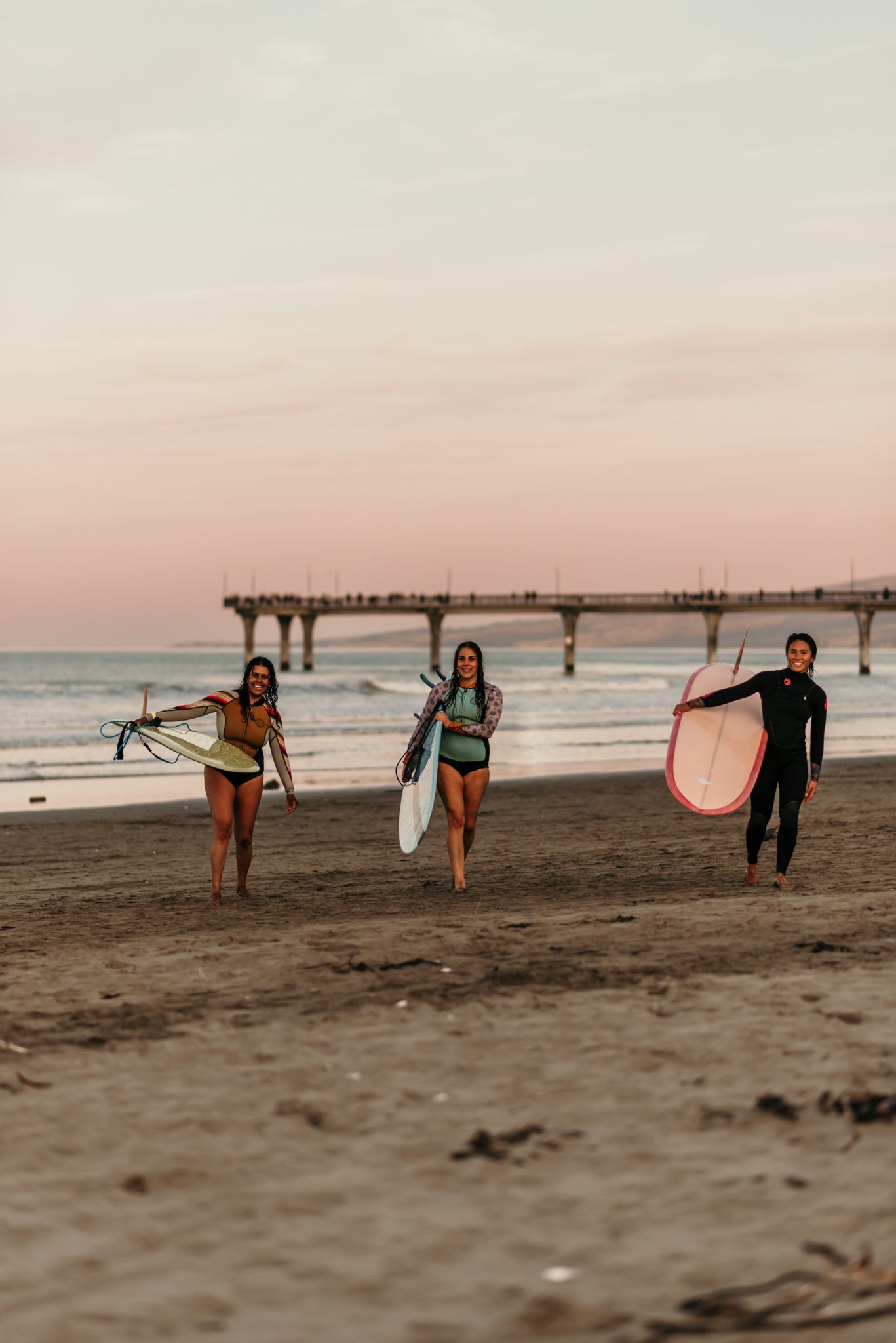 Three surfers walking along a beach with a pier in the distance during sunset.