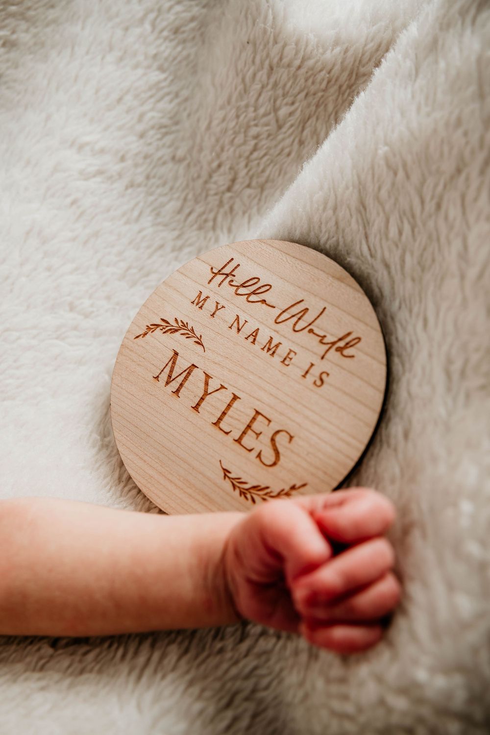 Wooden circular name plaque with 'Myles' engraved held by small hand against plush fabric.