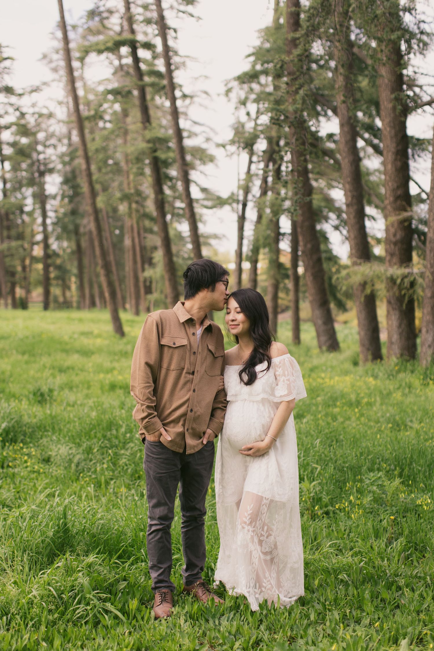 A couple poses together in a grassy field surrounded by tall evergreen trees.