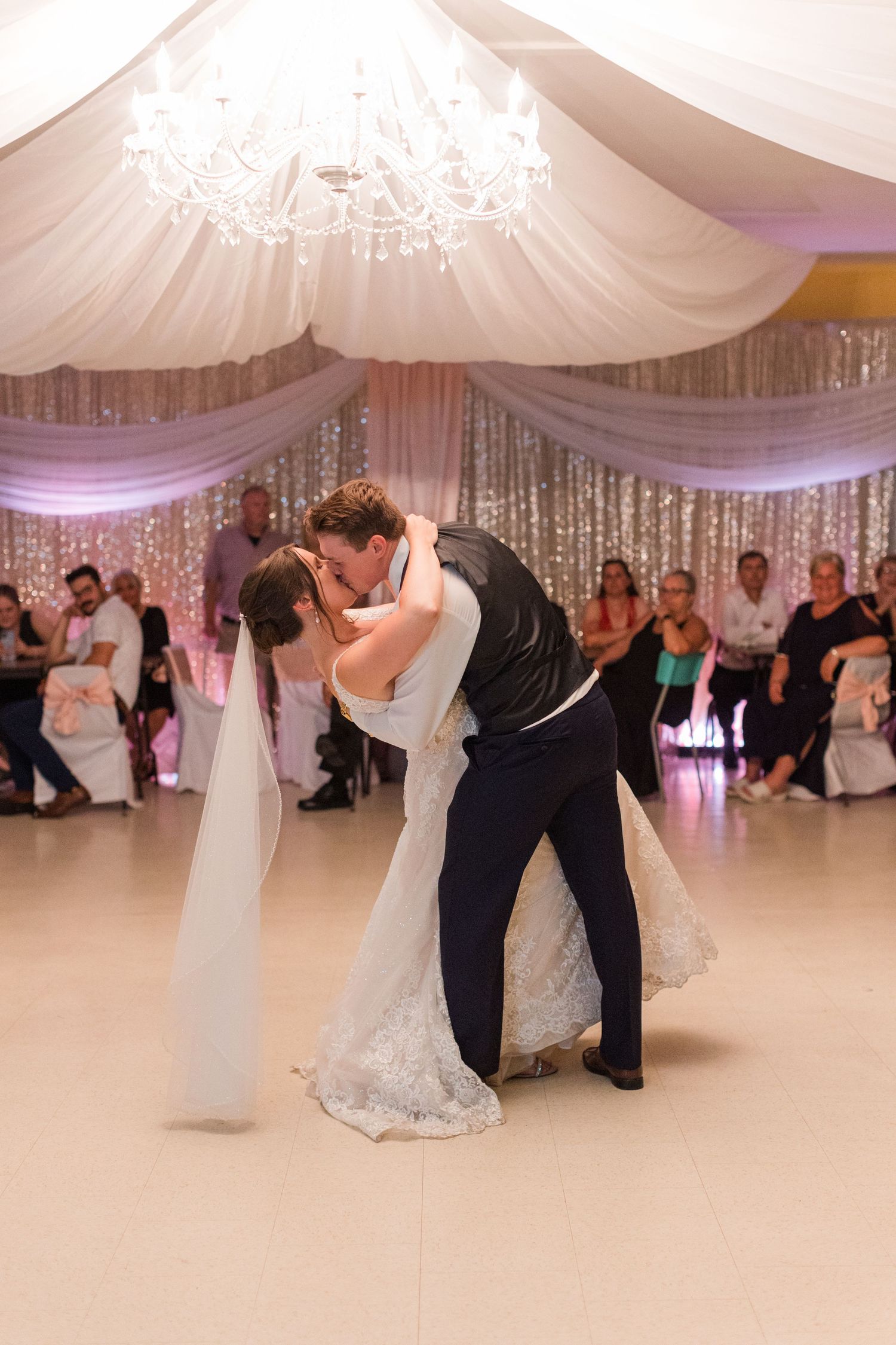 Couples dance gracefully at a wedding reception under elegant white draped ceiling and sparkling chandeliers.