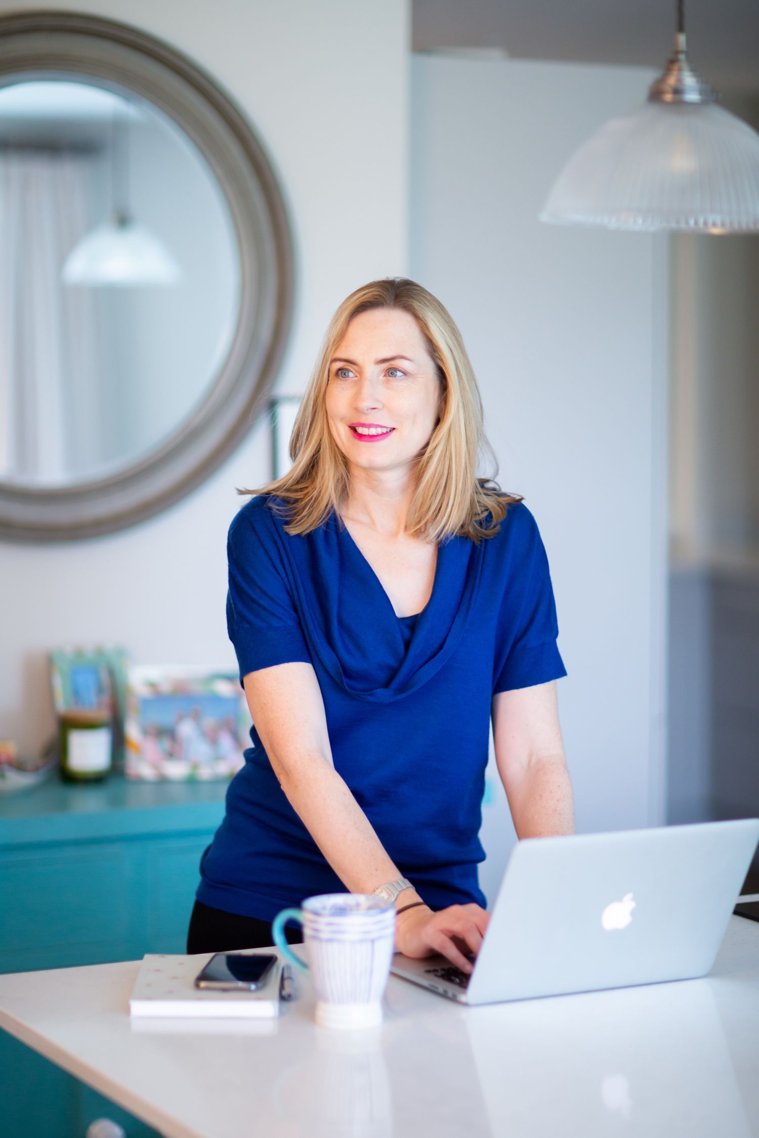 A professional works on a laptop at a desk with a round mirror and turquoise cabinet visible during business branding photoshoot.
