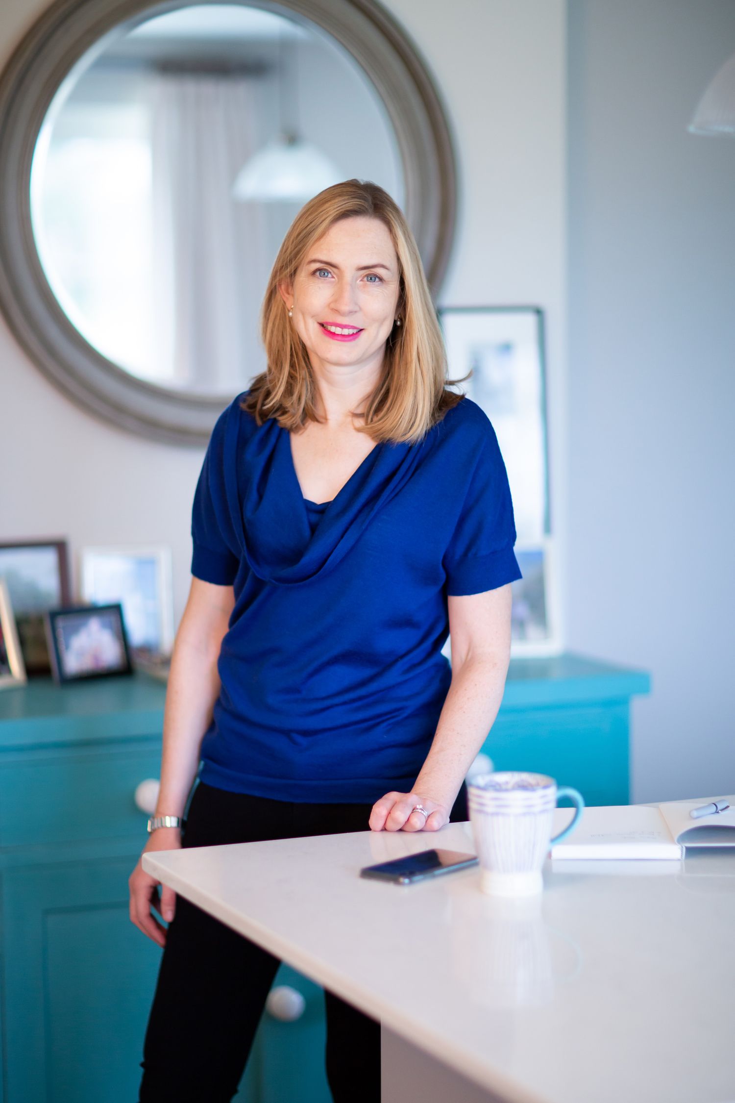 A professional stands at a desk wearing a blue cowl neck top near a round mirror during business branding photoshoot.