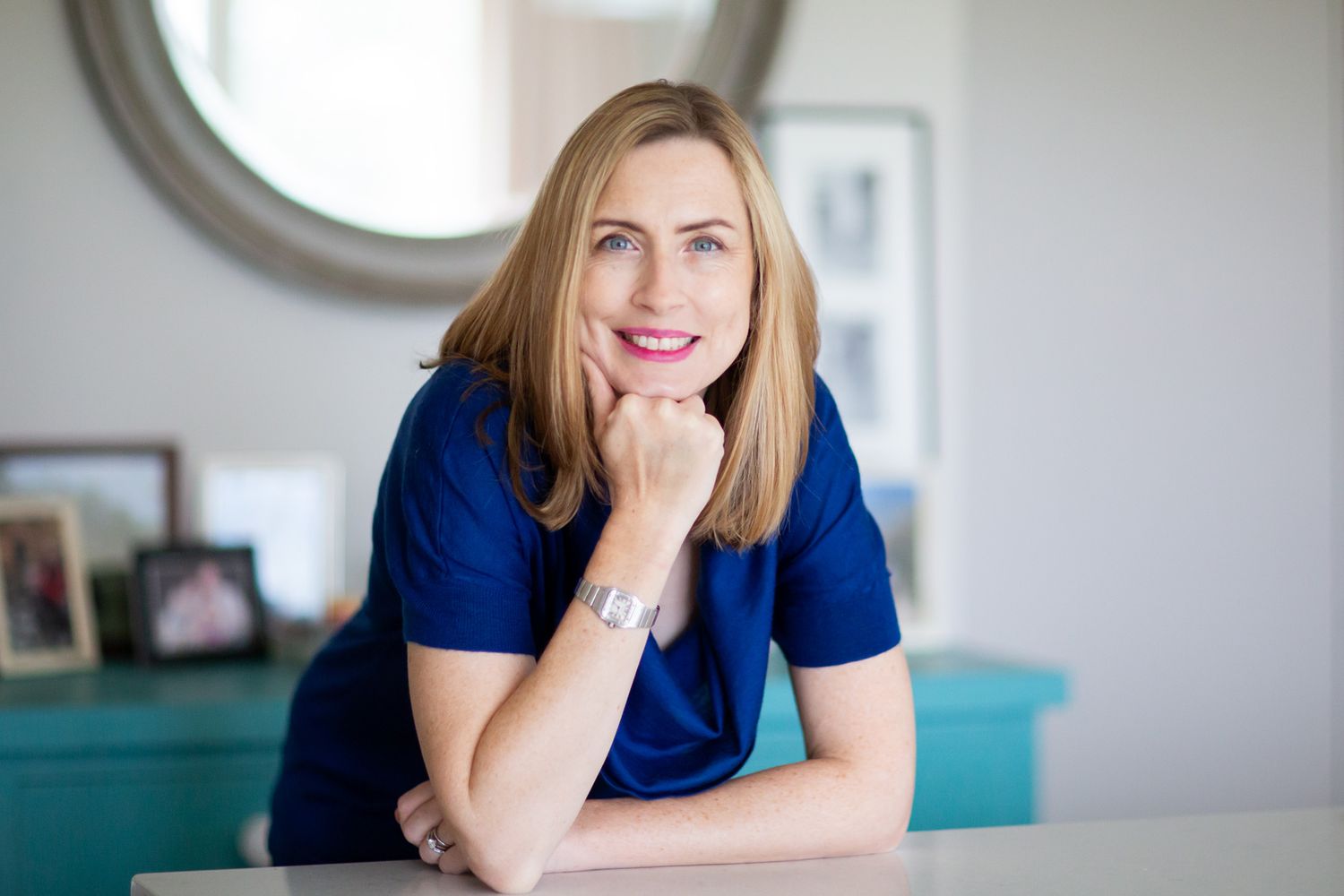 A professional poses casually at a desk with a turquoise cabinet and round mirror in background during business branding photoshoot.