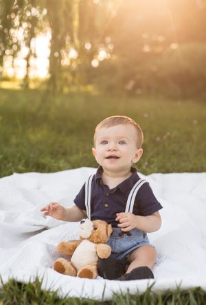 Baby wearing navy polo and suspenders sits on white blanket outdoors with teddy bear during golden hour sunset.