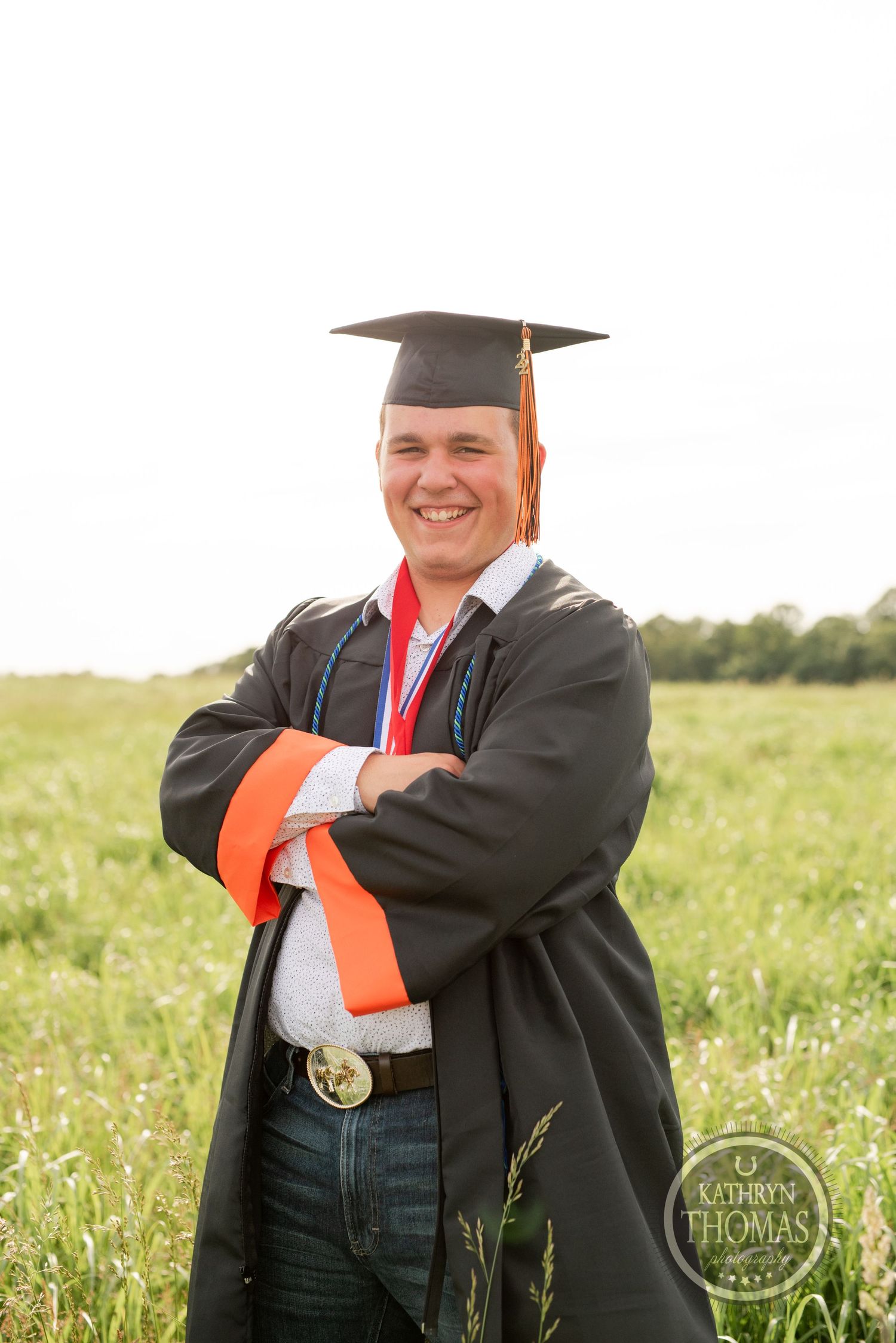 Graduate in black gown and orange stole poses in sunlit field during graduation.