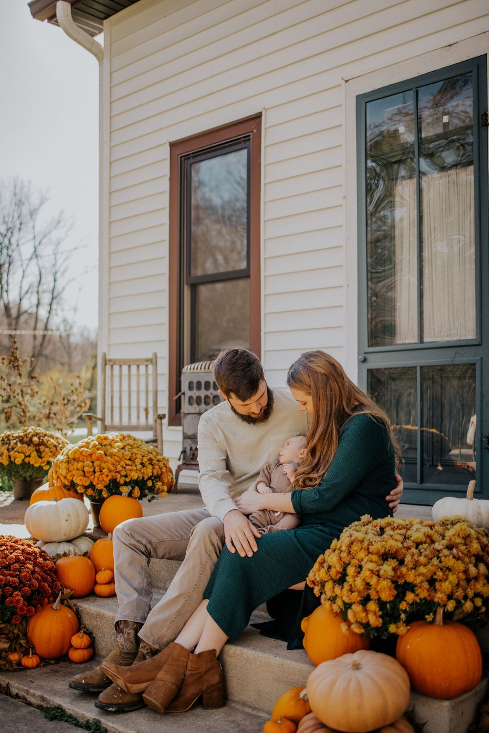 A couple sits together on a porch decorated with orange pumpkins and yellow mums for fall.