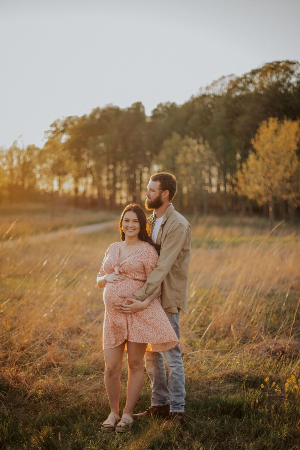 A couple embraces in a sunlit autumn field with tall golden grass and trees during a maternity photoshoot at sunset.