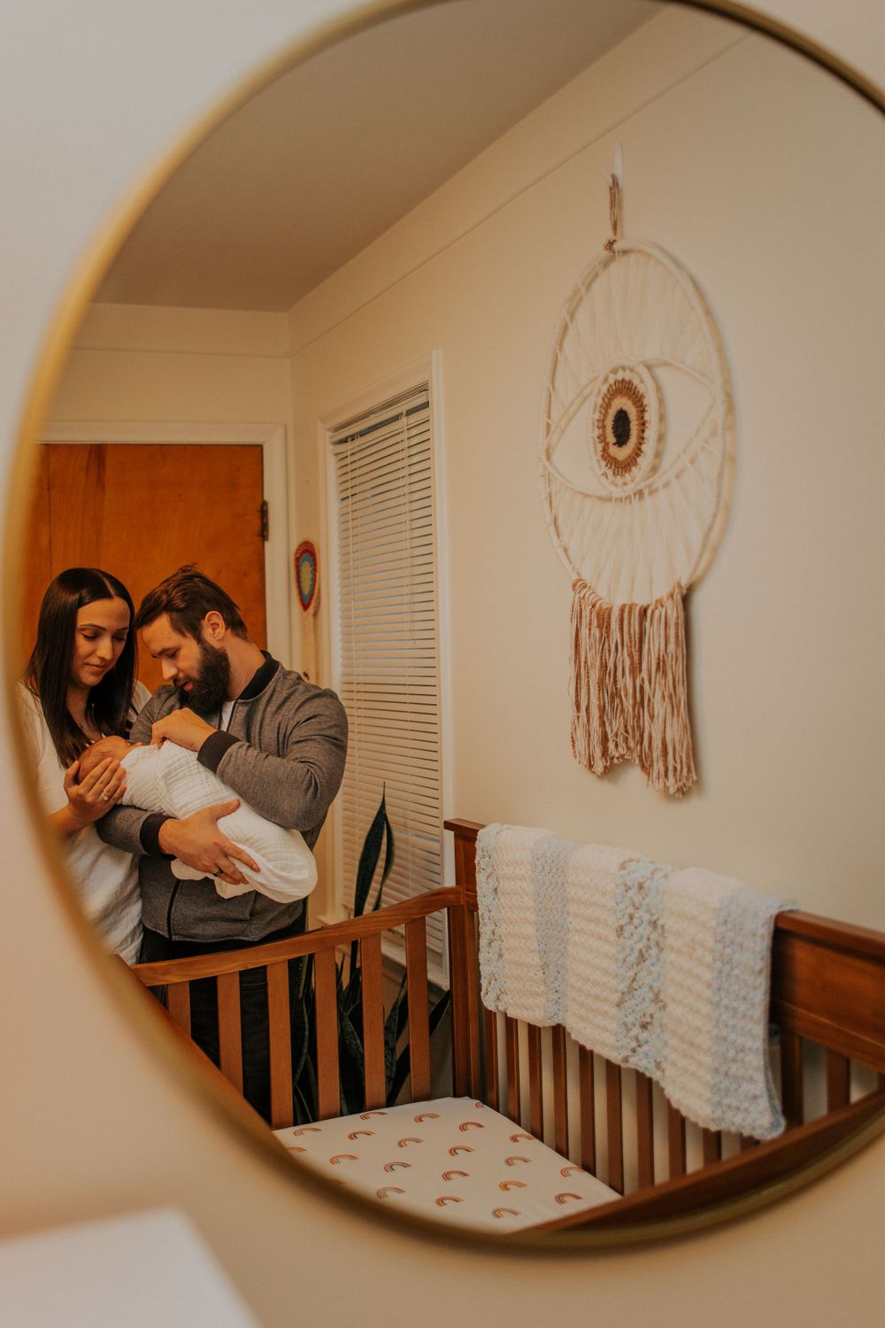 Mirror reflection shows couple with baby in nursery room with boho dreamcatcher wall decor and wooden crib.