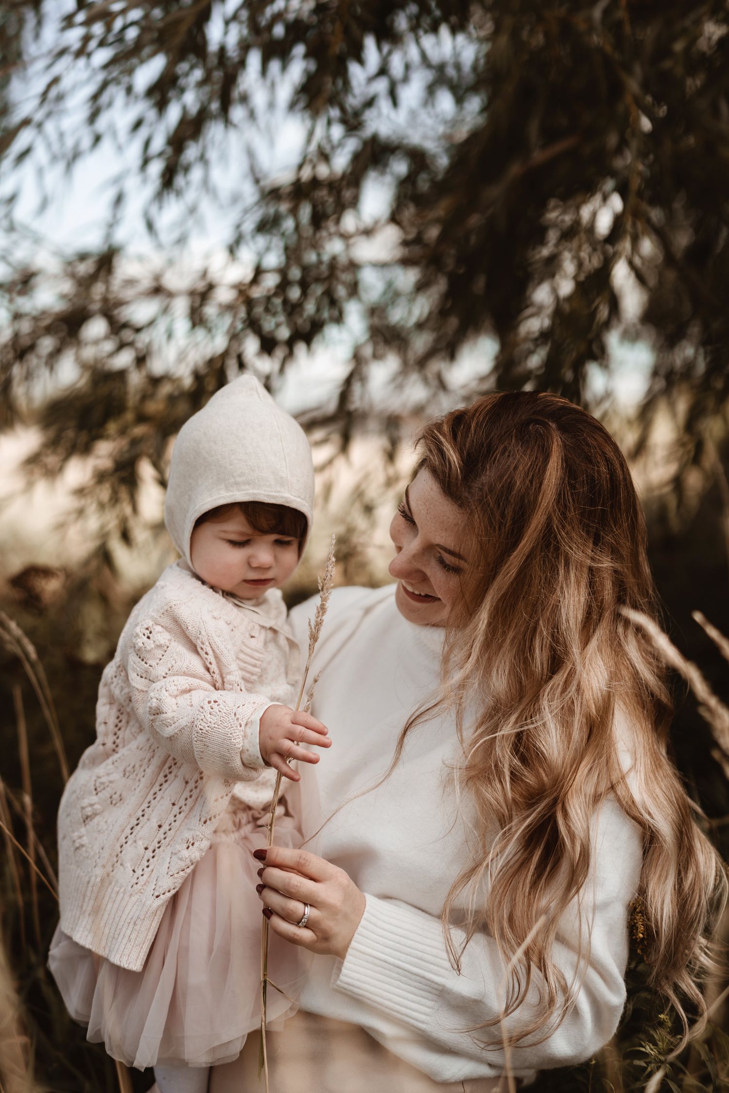 Tender outdoor portrait of parent and baby in white knit clothing under branches in warm autumn light.