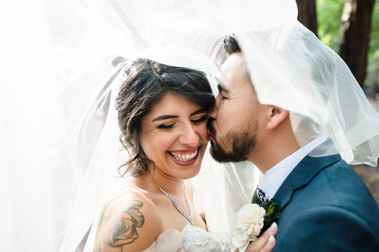 A couple shares a tender moment under a flowing white veil in a forest setting.