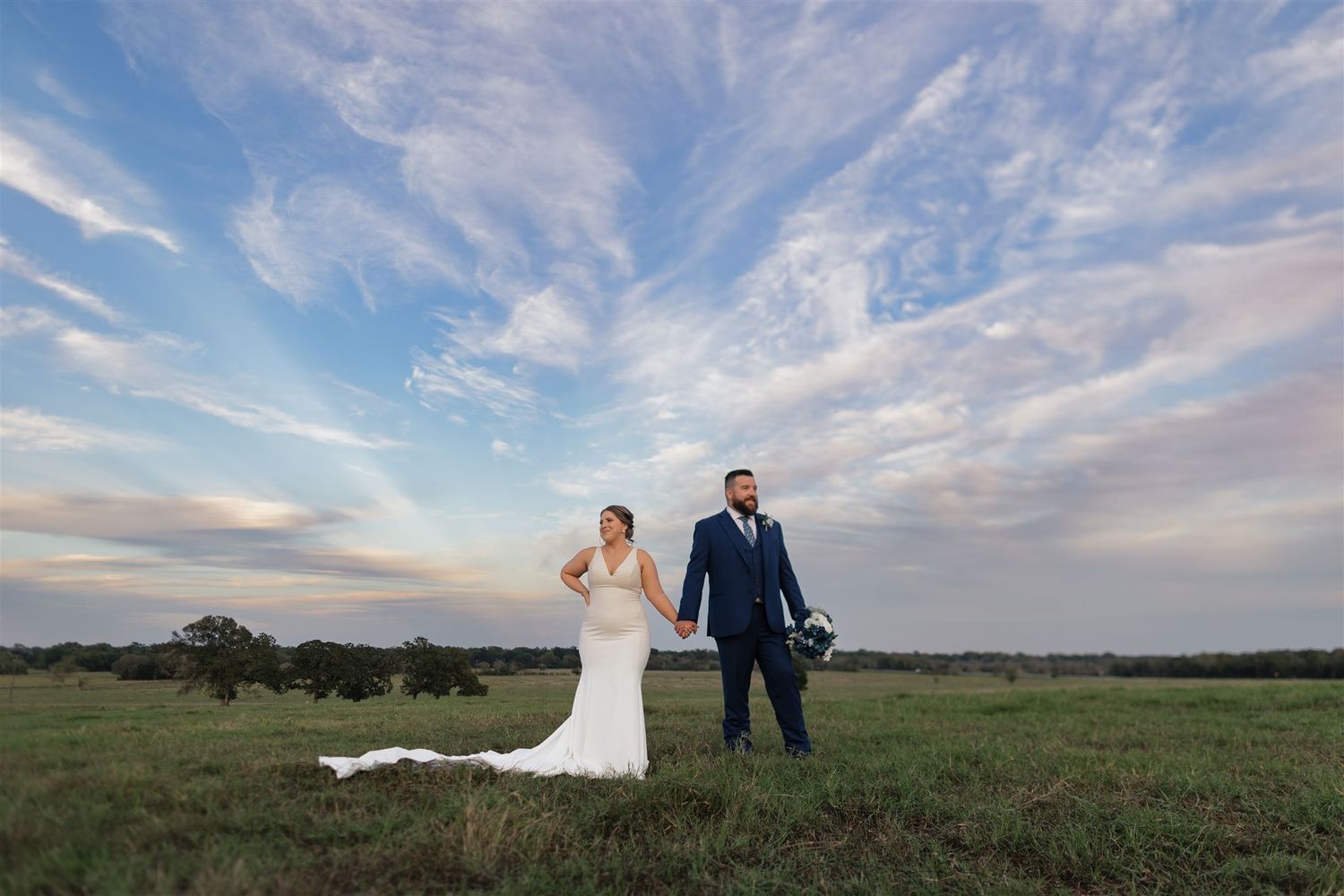 Newlyweds standing together in field under dramatic cloudy sunset sky.