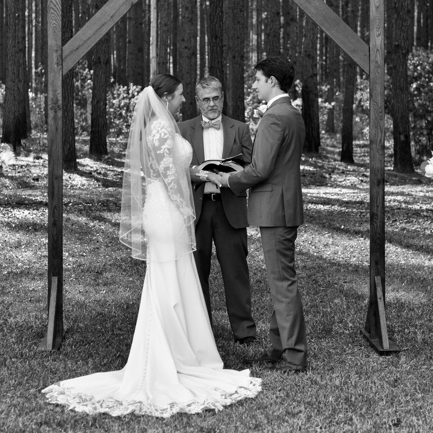 A wedding ceremony takes place under a wooden arch in a serene forest setting with tall pine trees in the background.