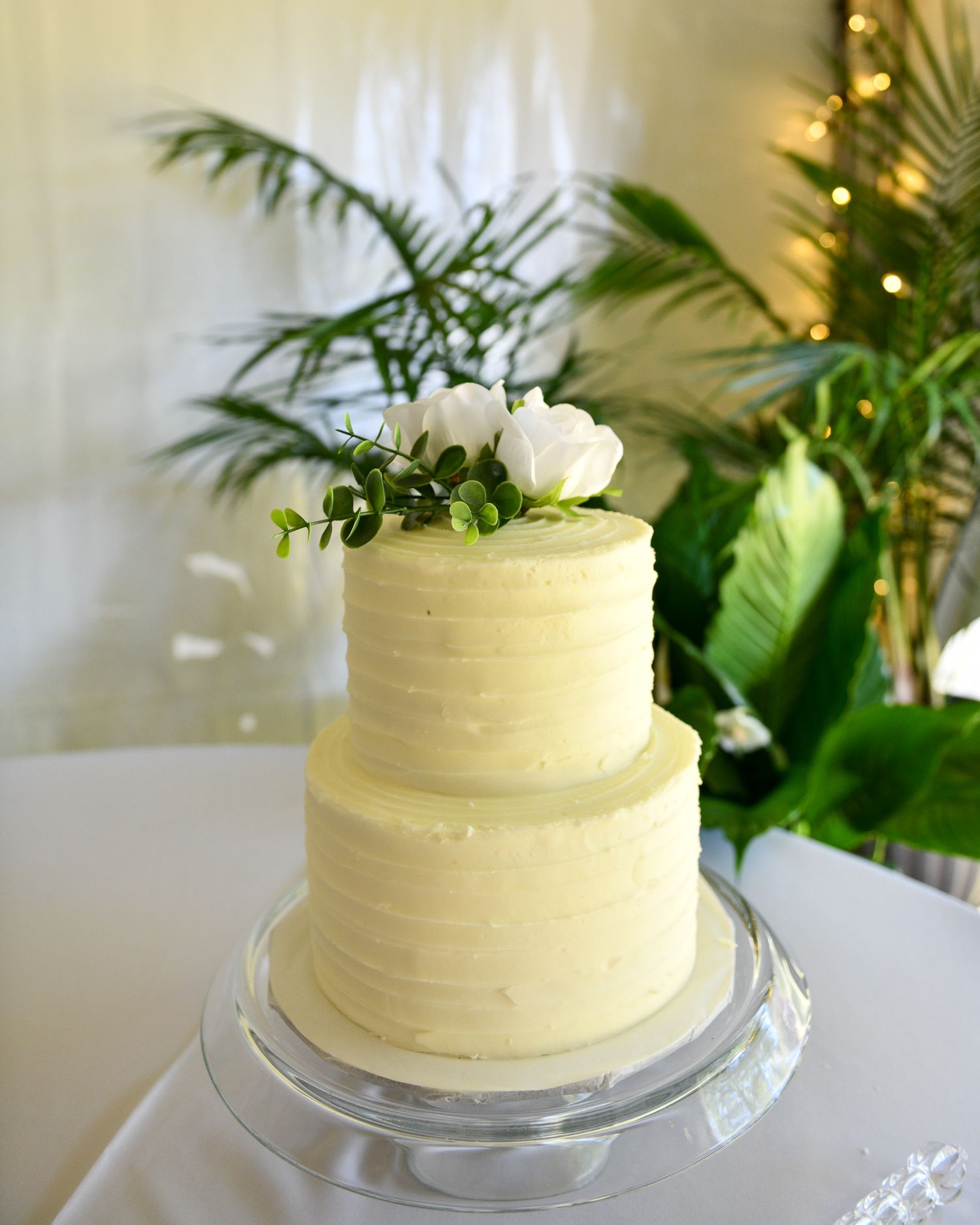 A two-tier cream colored wedding cake decorated with white flowers and greenery on a crystal stand.
