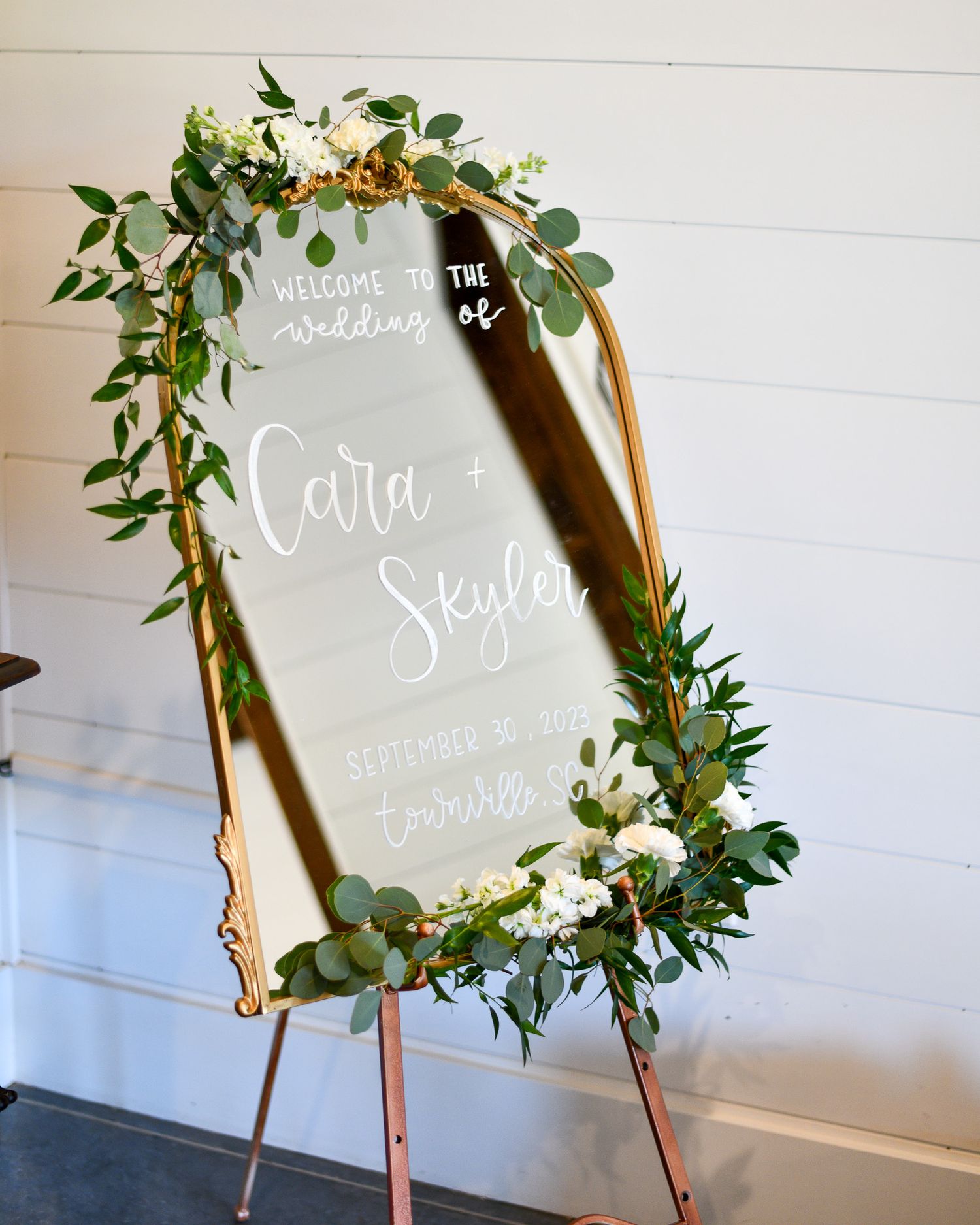Elegant wedding welcome sign with greenery and floral decorations on a gold-framed mirror.