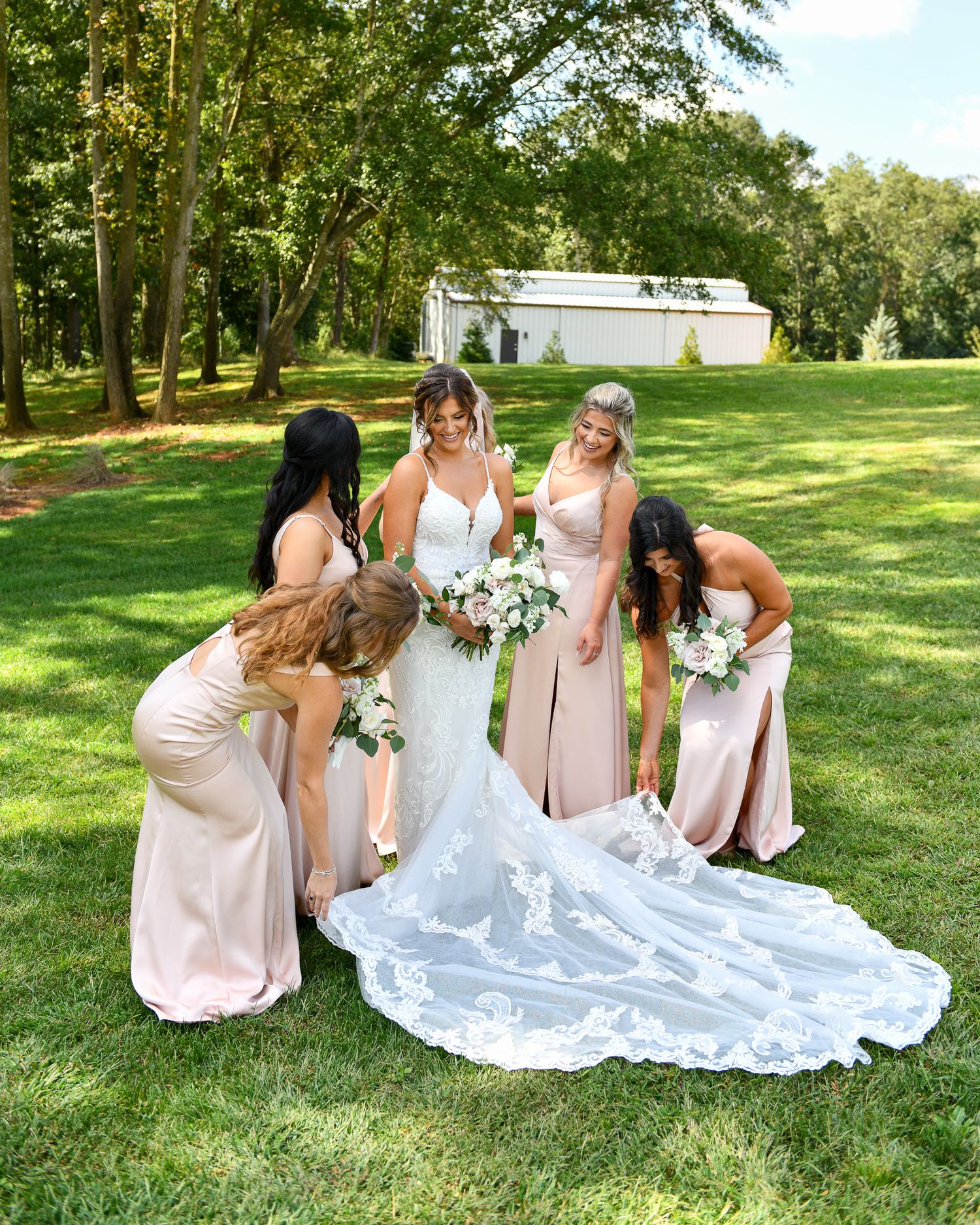 A bridal party in pink dresses arranges the train of a lace wedding gown on a sunny lawn.