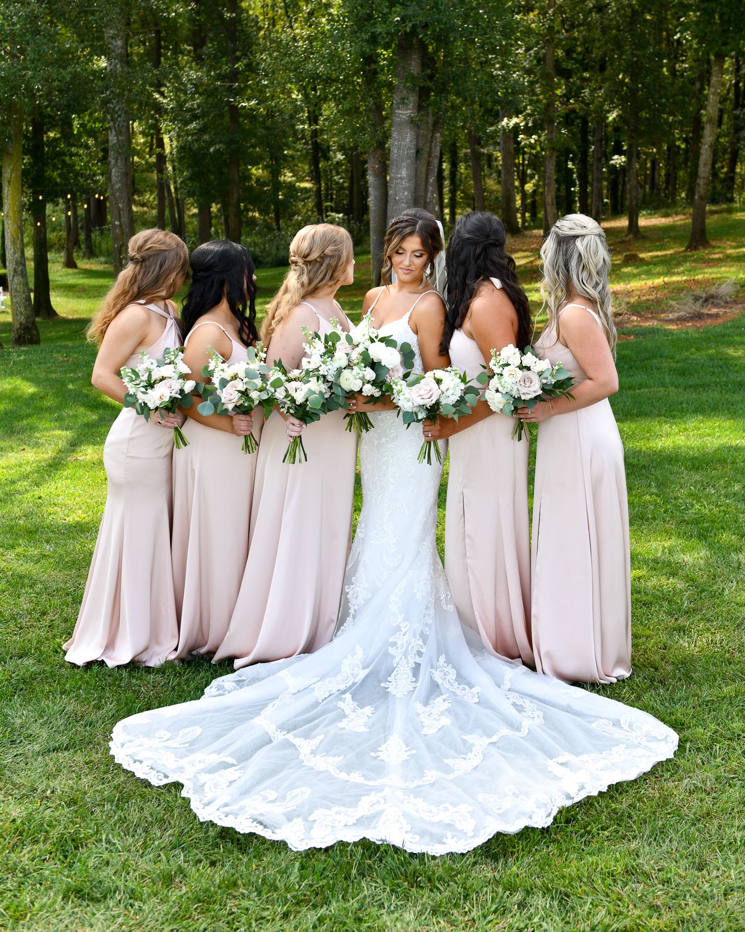 A bridal party wearing blush pink dresses poses in a garden setting with white floral bouquets.