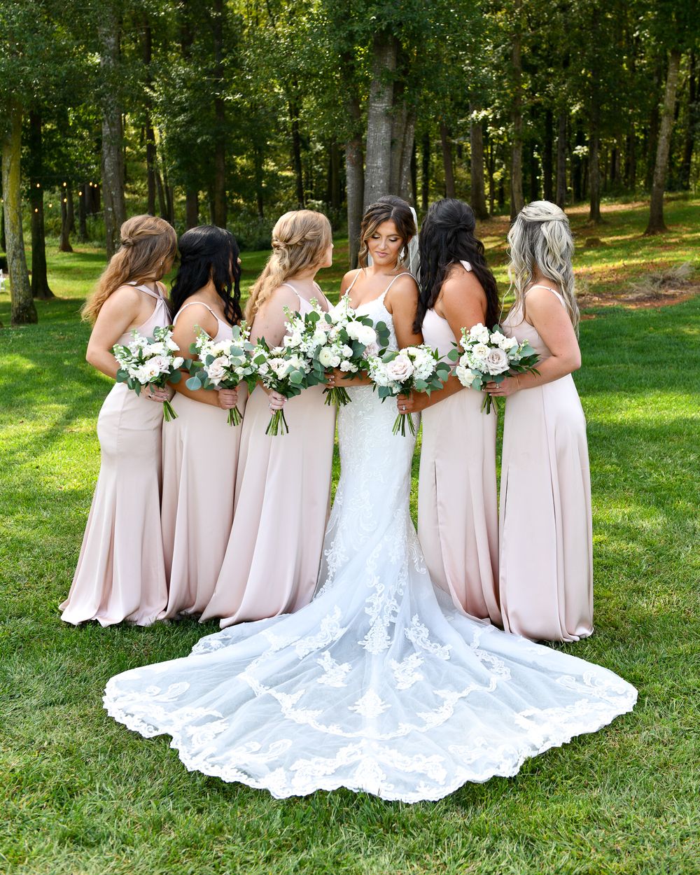 A bridal party wearing blush pink dresses poses in a garden setting with white floral bouquets.