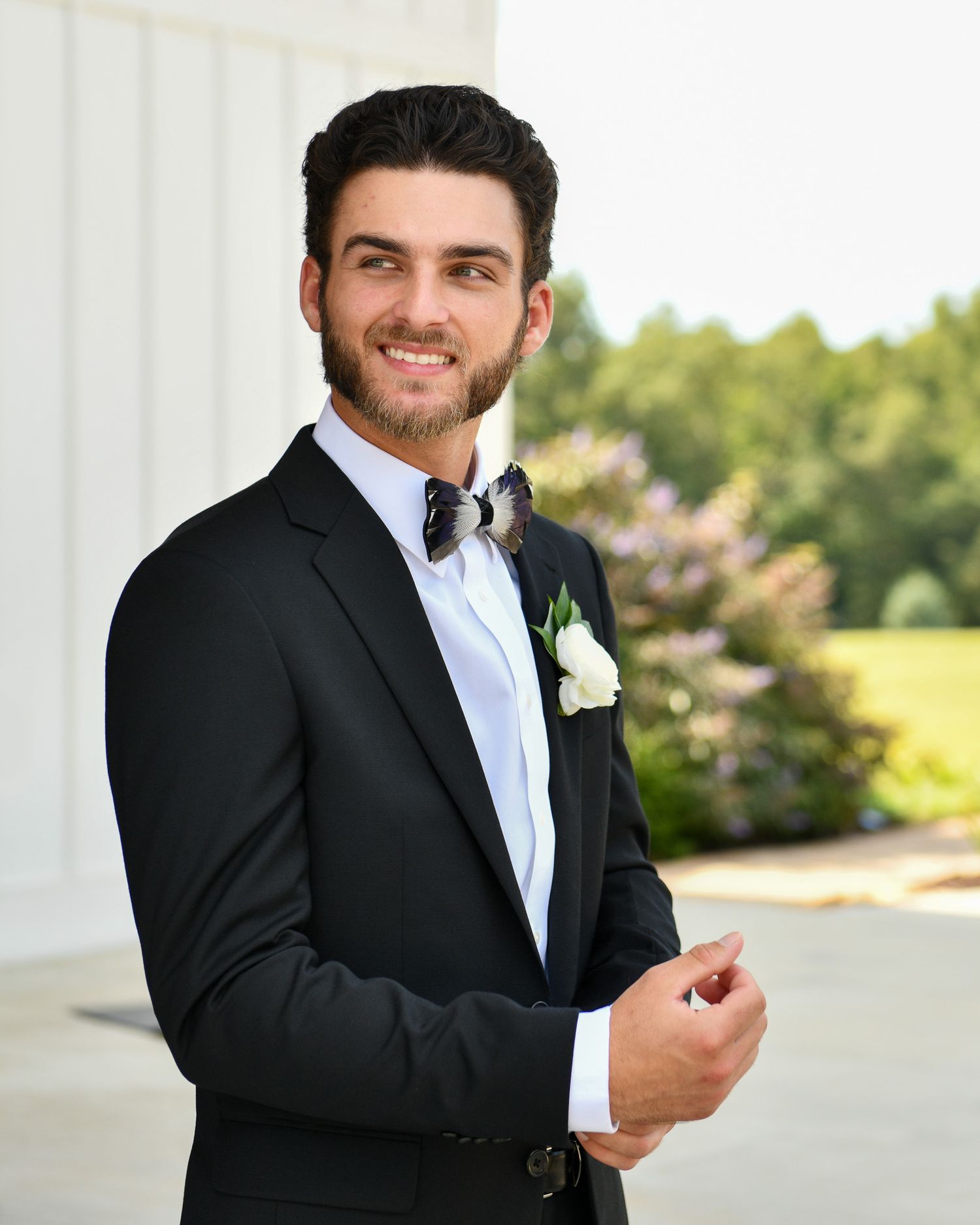 A groom in classic black tuxedo with white boutonniere poses on a white columned porch.