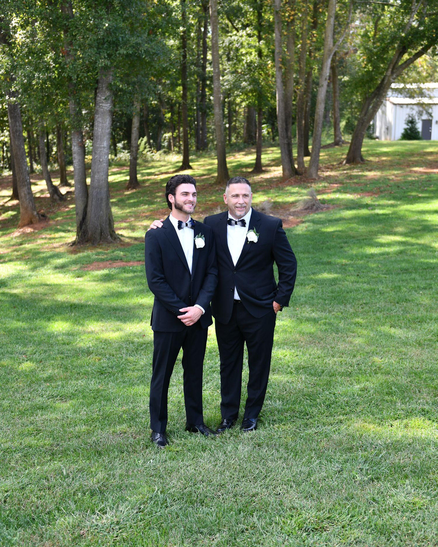 Two gentlemen in black tuxedos stand together on a wooded lawn.