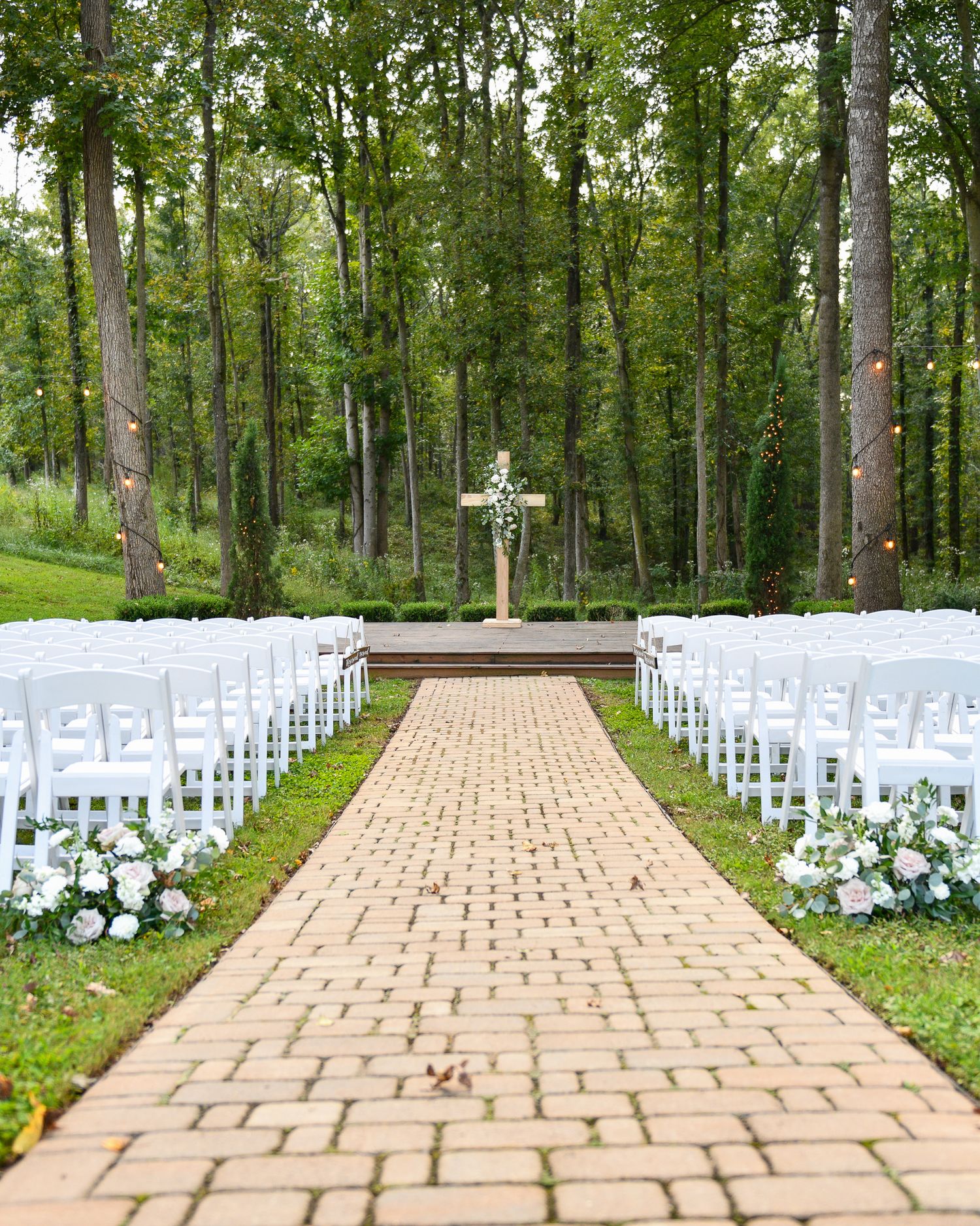 A beautiful outdoor wedding ceremony setup with white chairs and a brick aisle leading to a cross in the woods.