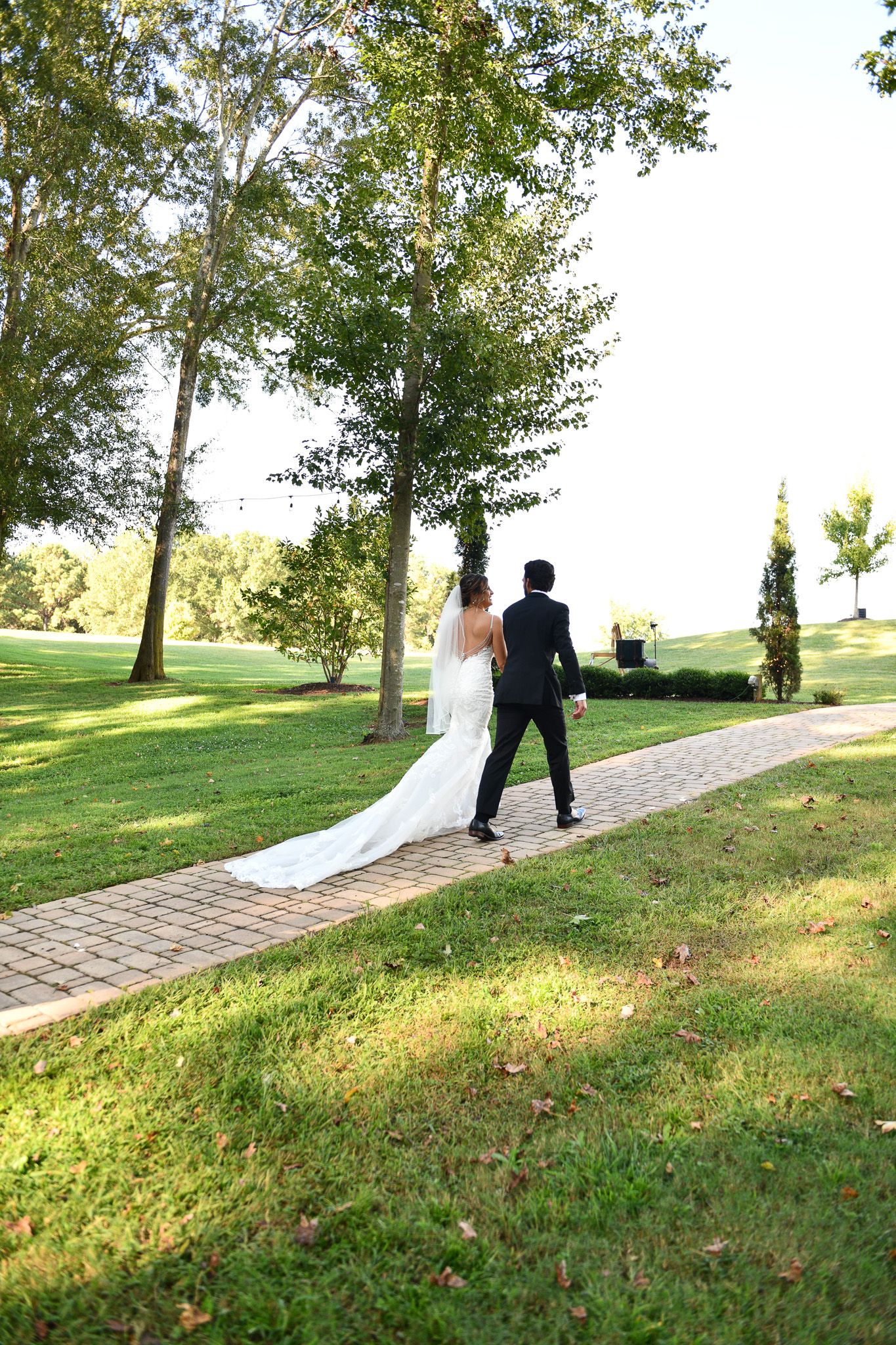 A couple walks down a brick path surrounded by trees at their outdoor wedding venue.