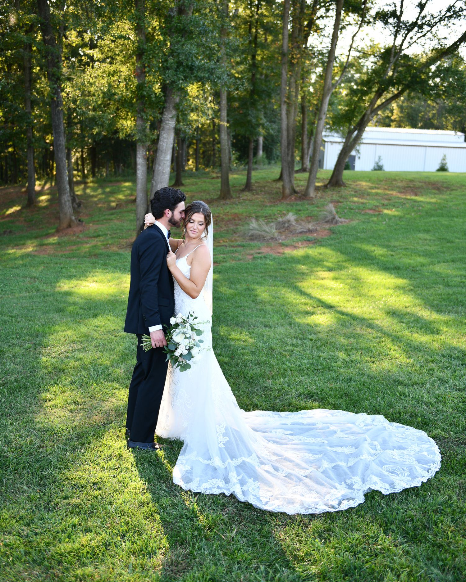 A romantic wedding portrait in a wooded setting with a dramatic lace train spread on green grass.