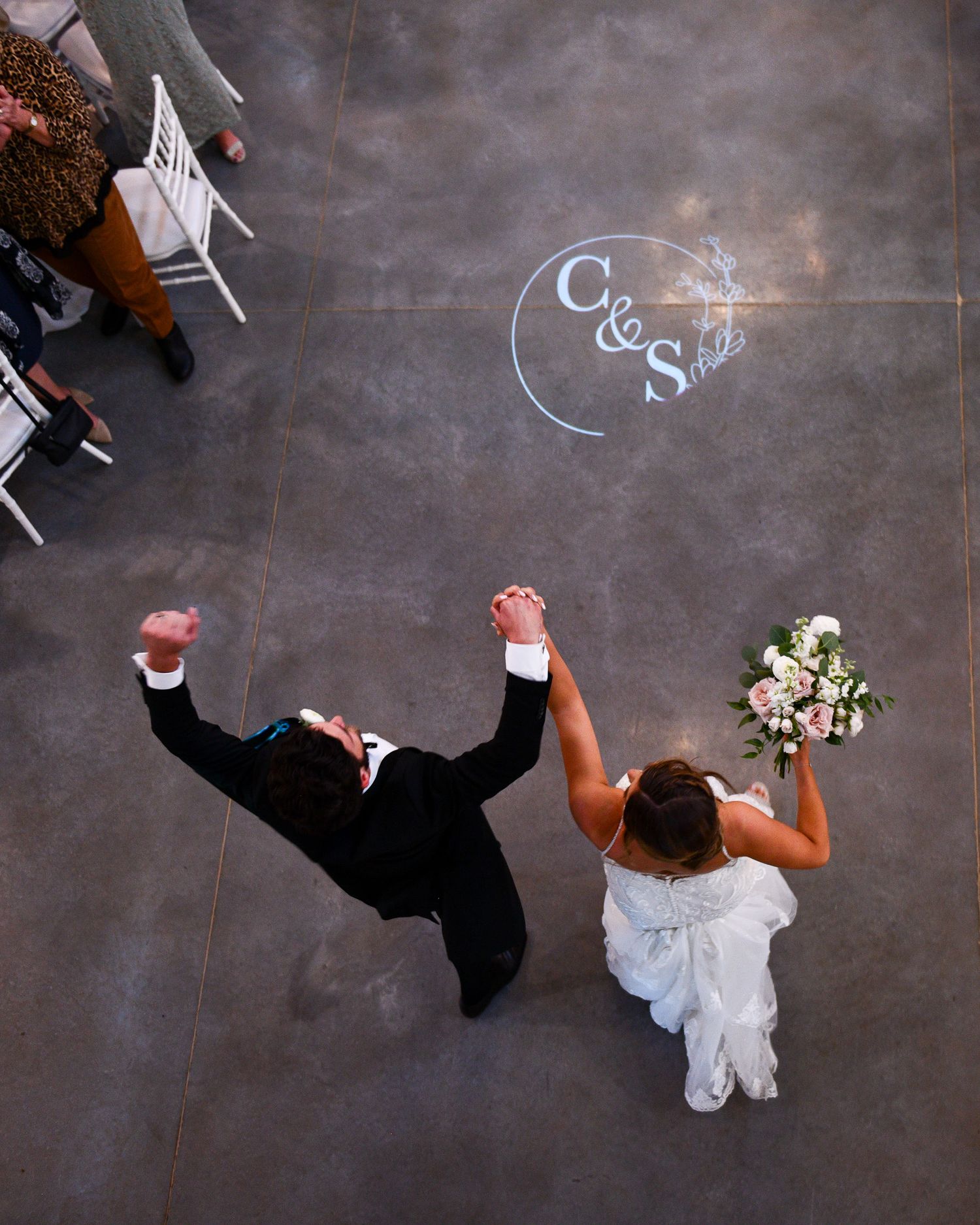 Aerial view of newlyweds celebrating with raised arms on a floor with illuminated monogram.