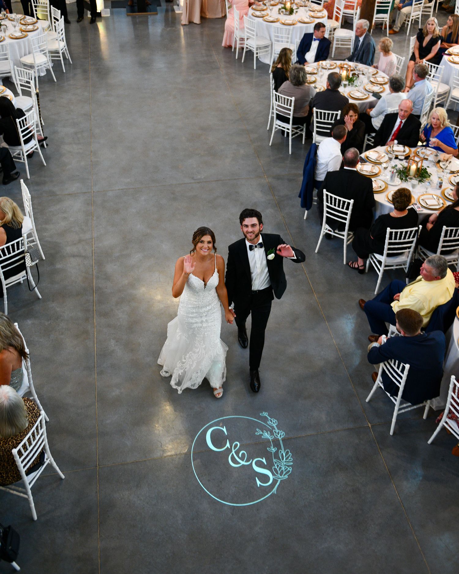Overhead view of a reception venue with white chairs and a glowing monogram on concrete floor.