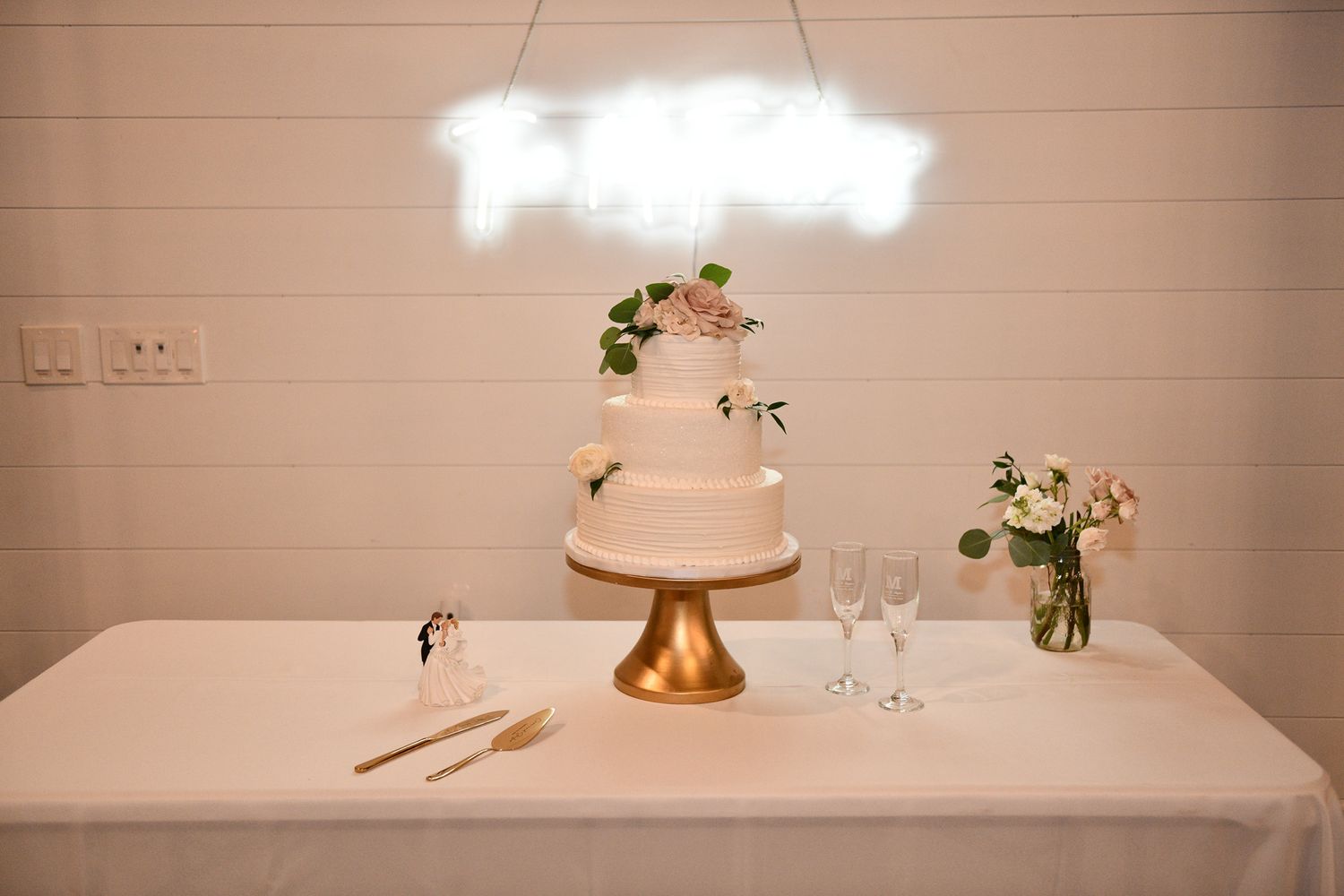 Elegant white wedding cake with floral decorations displayed on a gold stand with a neon sign above.
