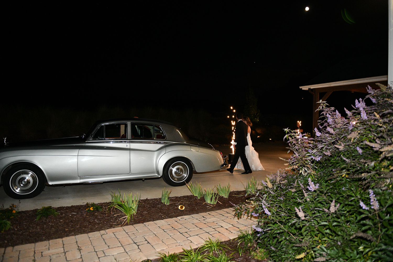Classic silver vintage car parked near illuminated garden pathway at night.