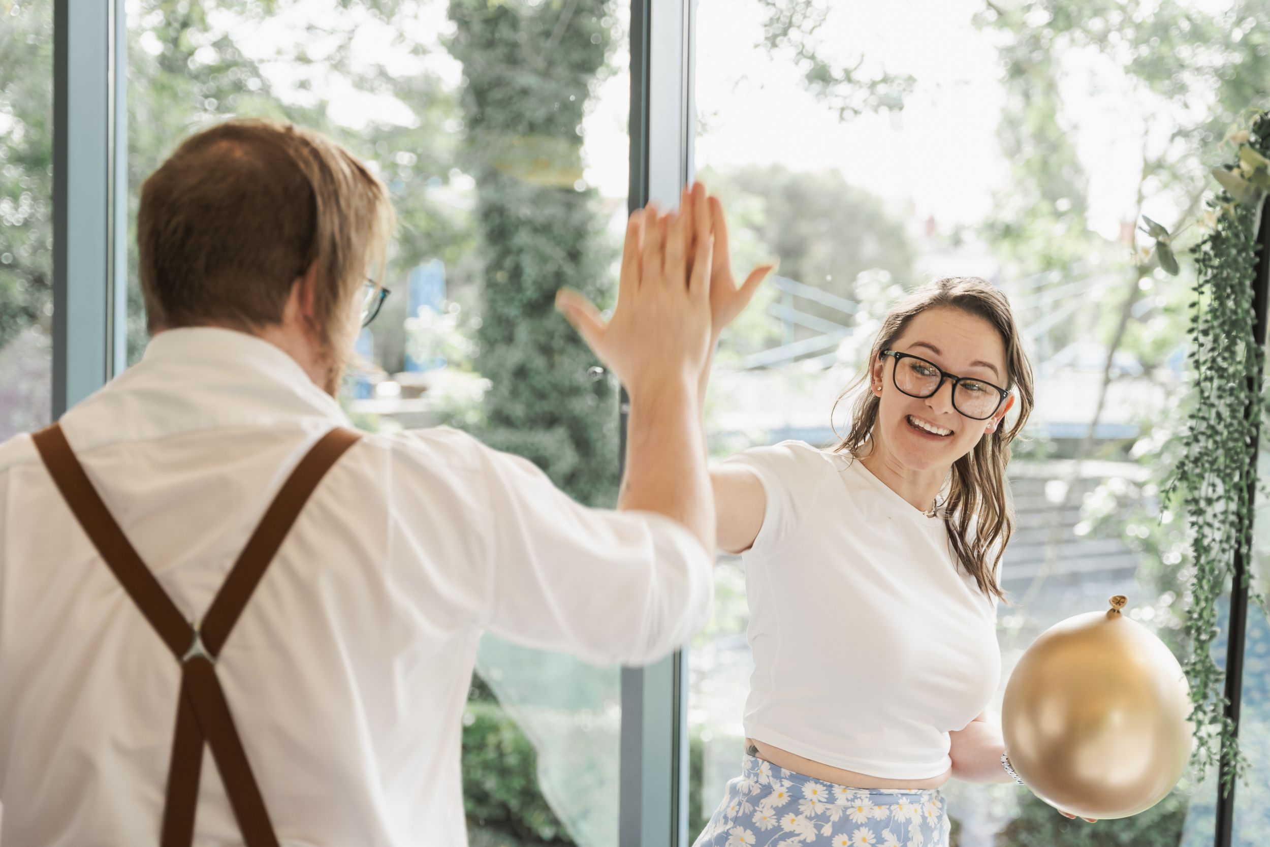 A joyful couple celebrating with raised hands and gold balloons in a bright sunlit room.