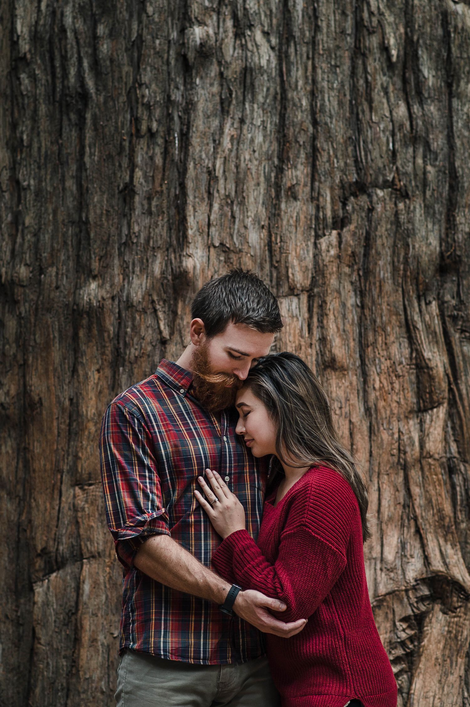 A couple embraces tenderly against a rustic wooden backdrop with red and plaid attire.