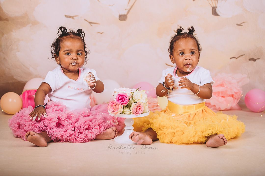 Twin babies in white outfits with pink and yellow tutus sitting with birthday cake.