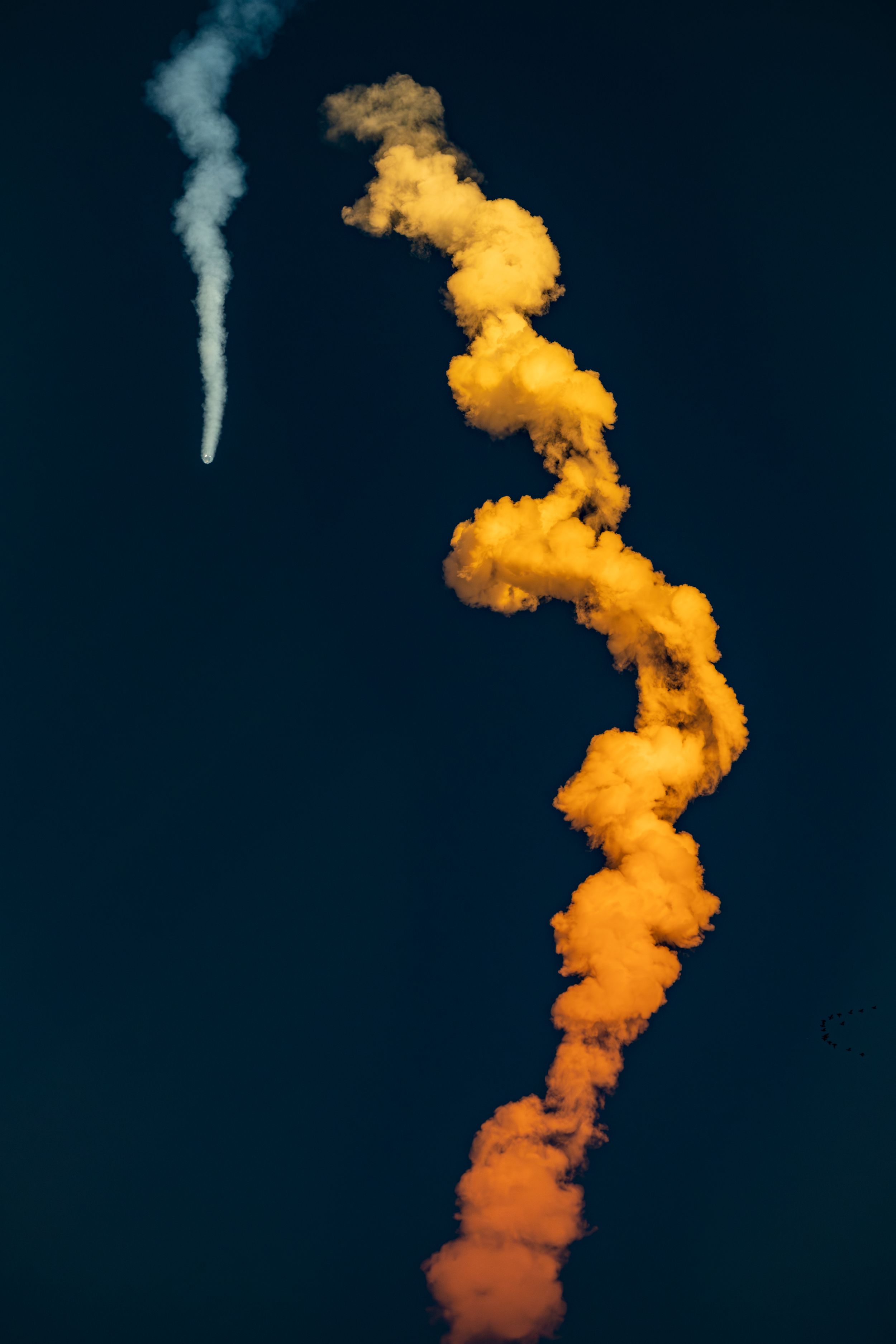 Orange rocket exhaust trail curling against deep blue sky with white smoke streak.