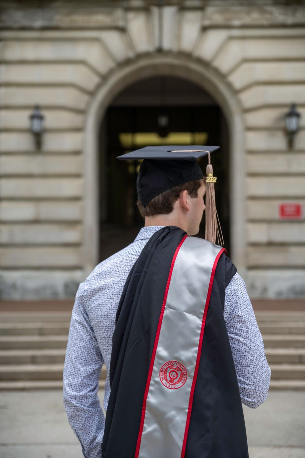 Graduate wearing black cap and gown with decorative white and red sash seen from behind.