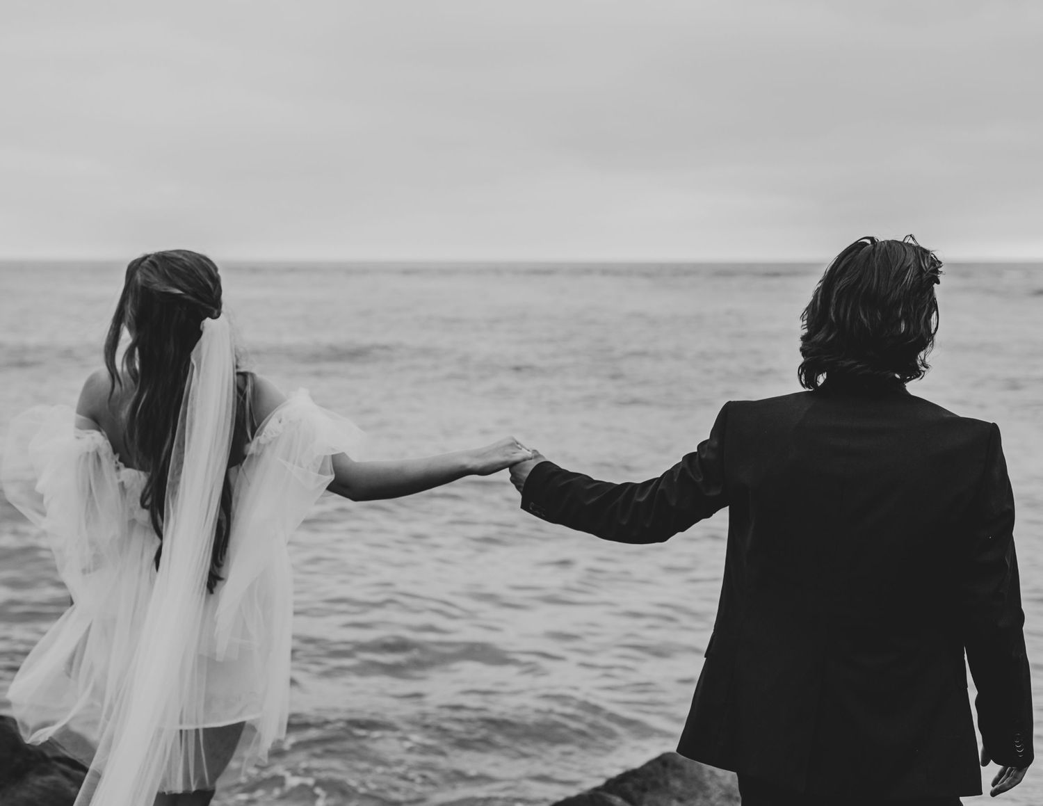 Black and white photo of newlyweds holding hands by the ocean.