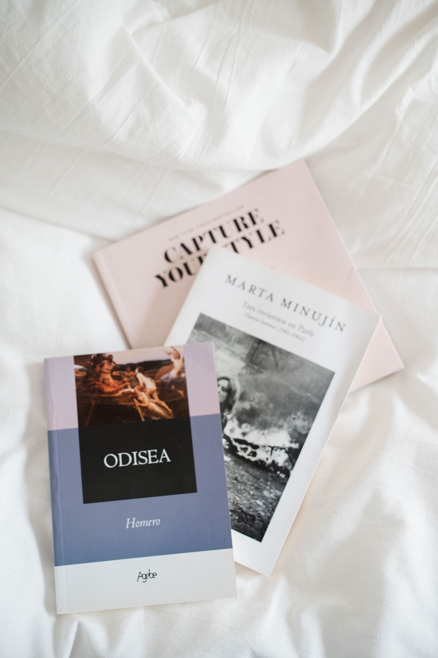 Three books including Odisea lay on white fabric in an artistic arrangement.
