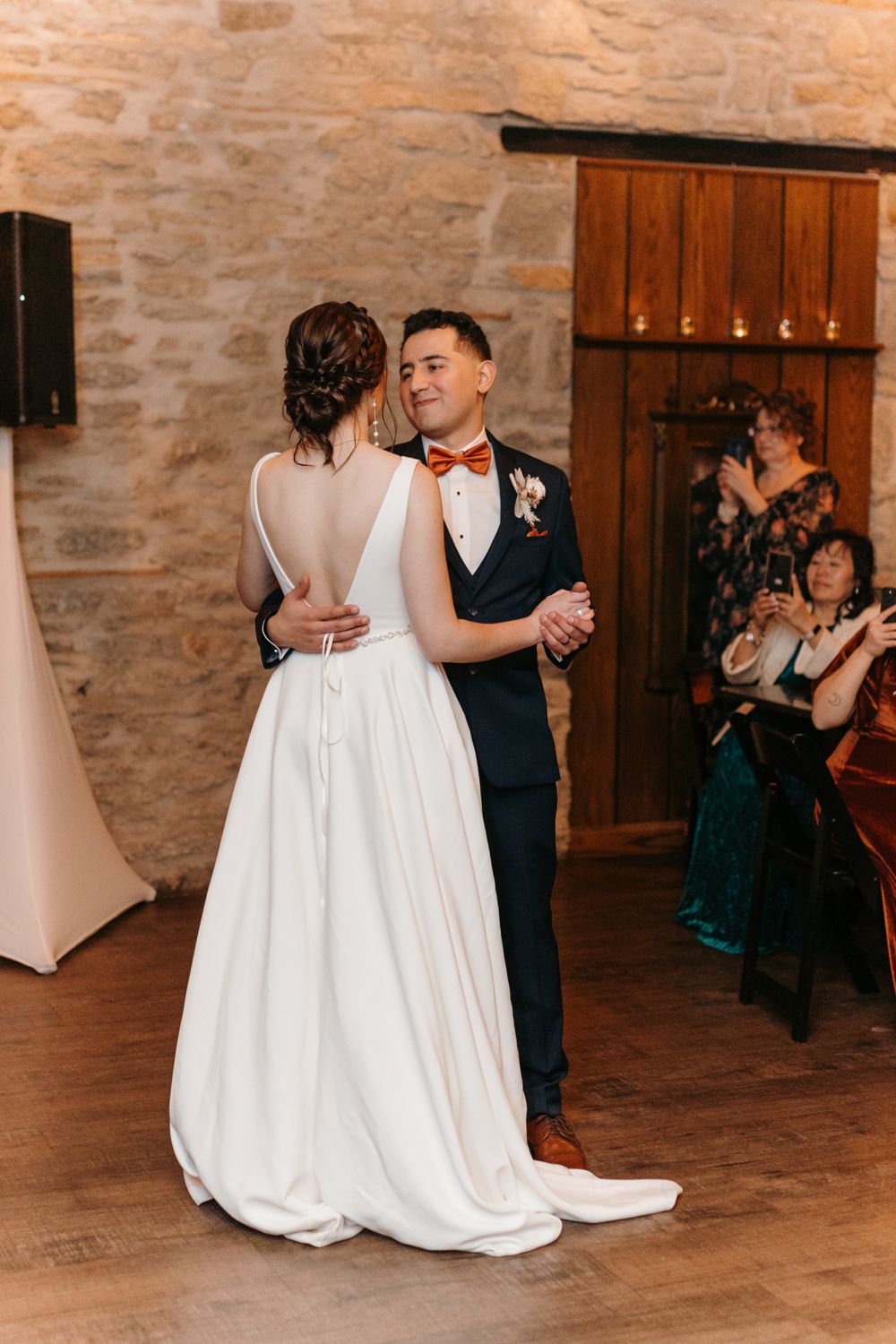 Groom in navy suit with orange bow tie shares first dance with bride in a backless white gown.