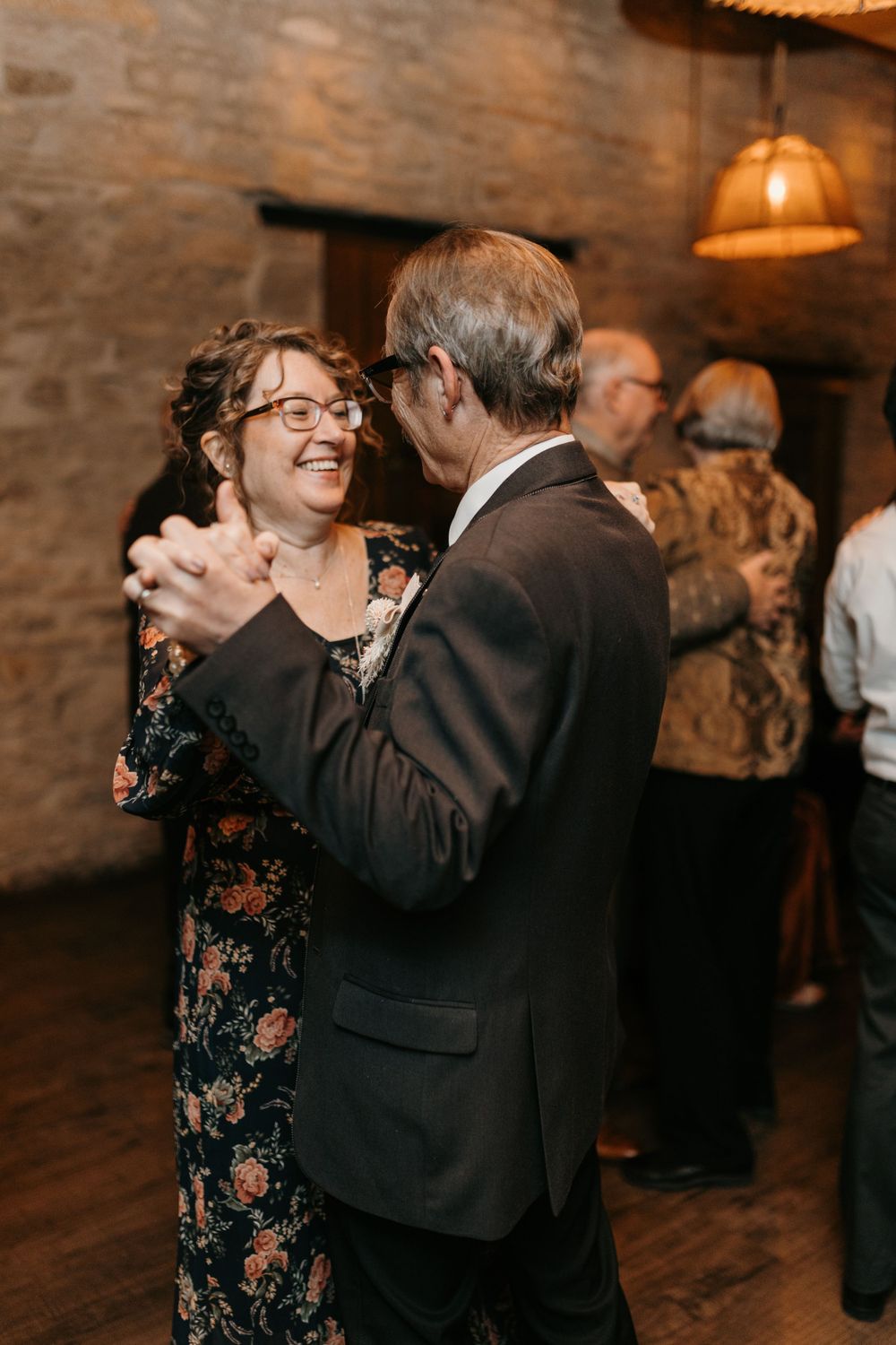 Joyful woman in floral dress dances with a man in a dark suit at a rustic stone wedding venue.