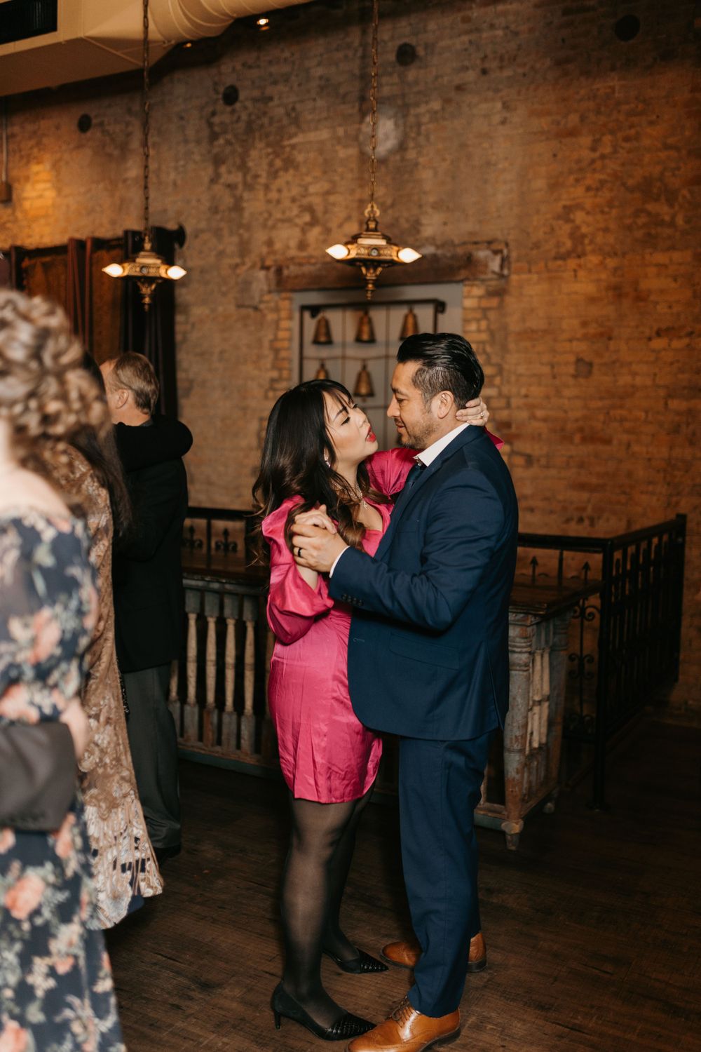 A couple in formal attire shares an intimate dance at a wedding reception in a brick-walled venue.