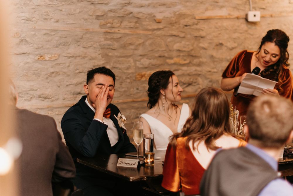 Emotional groom reacting to a wedding speech while bride smiles at rustic reception.