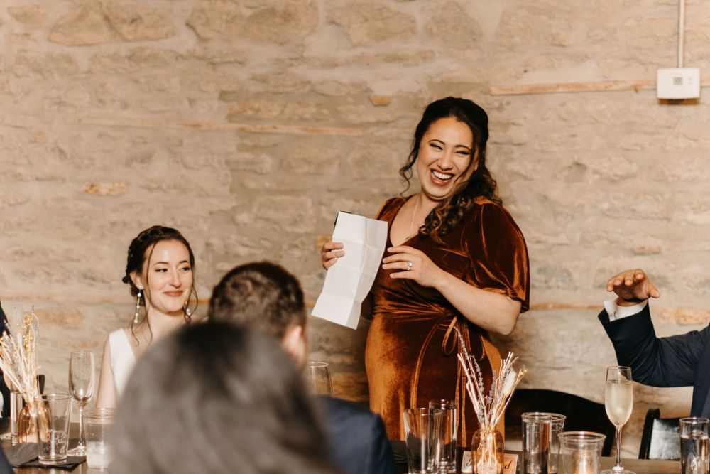 Bridesmaid laughing while giving a toast at an intimate rustic stone wedding reception.