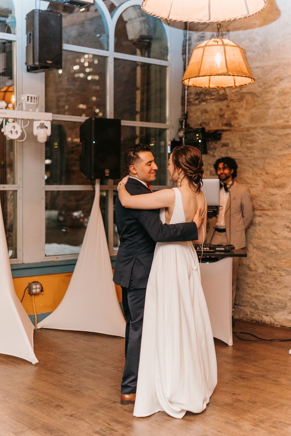 Bride and groom share a romantic first dance under a vintage lamp with DJ visible in background.