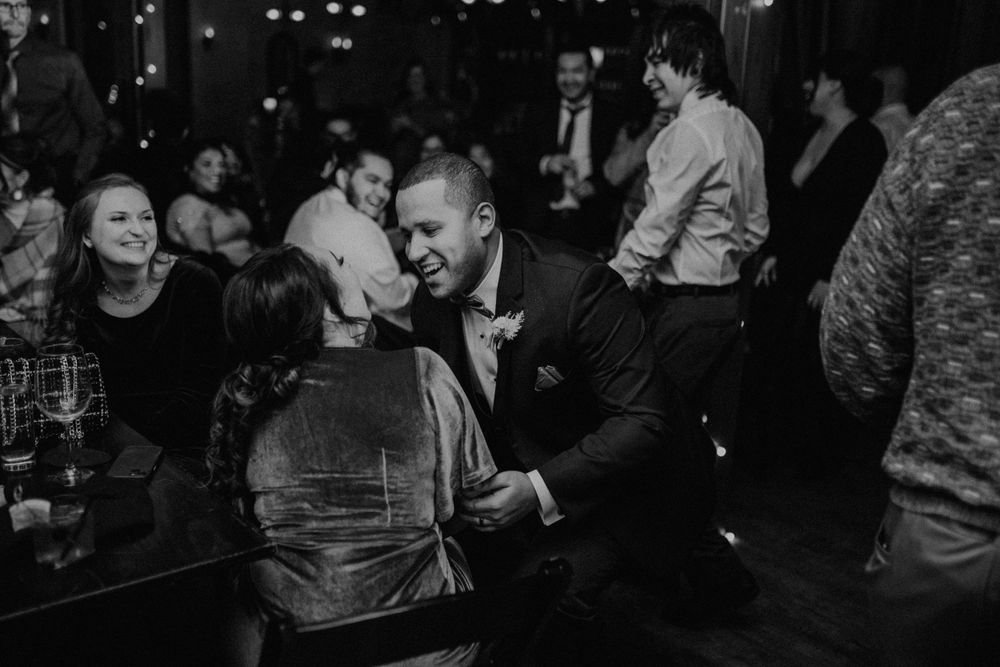 Groom in tuxedo shares intimate moment with guest at wedding reception in black and white photo.