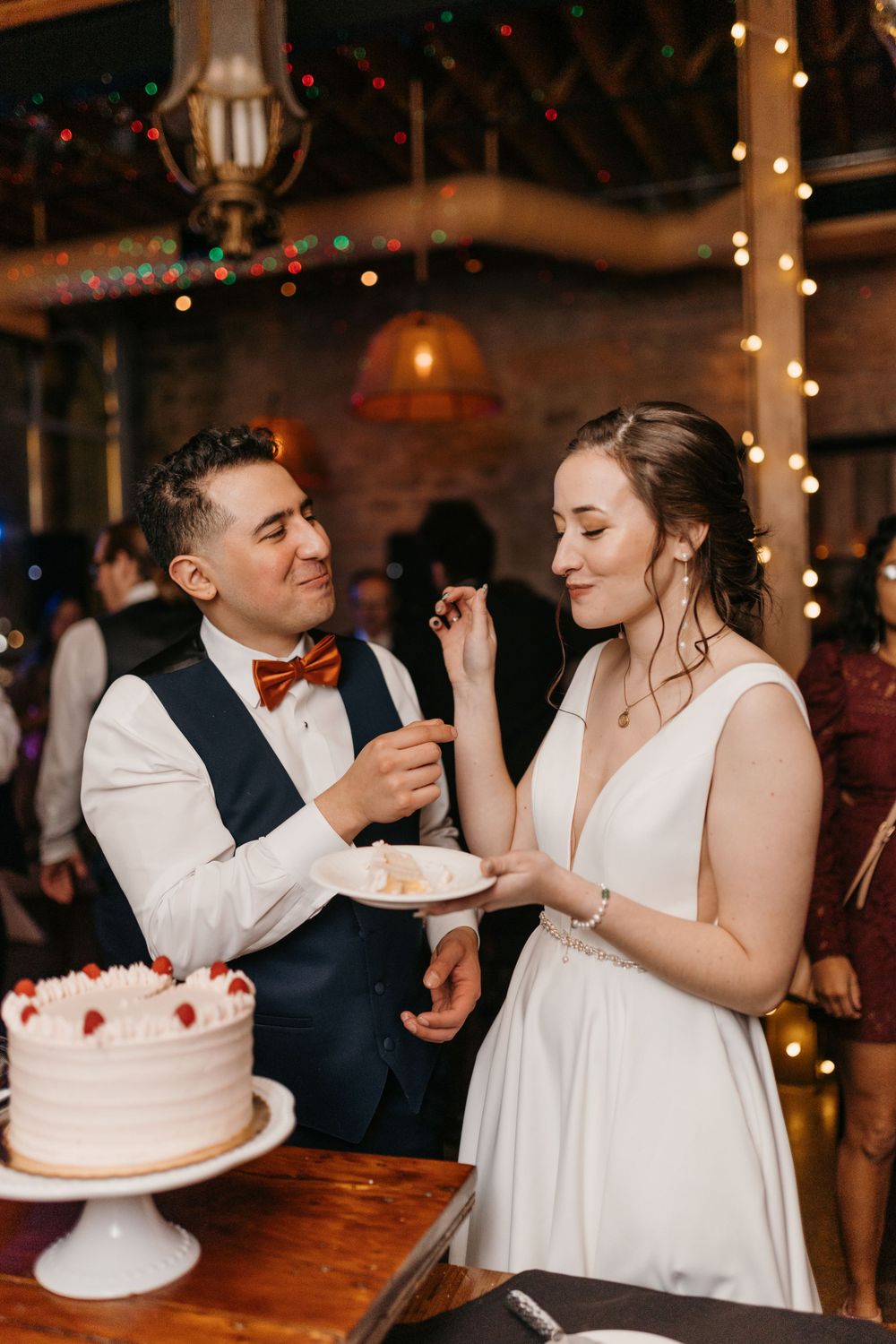 Newlyweds sharing wedding cake cutting moment at elegant reception with warm string lights.