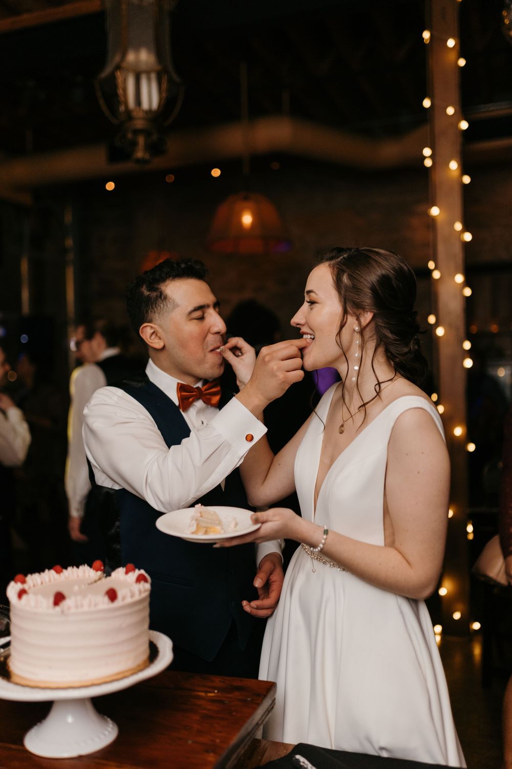 Bride and groom feeding each other wedding cake at romantic reception with string lights.