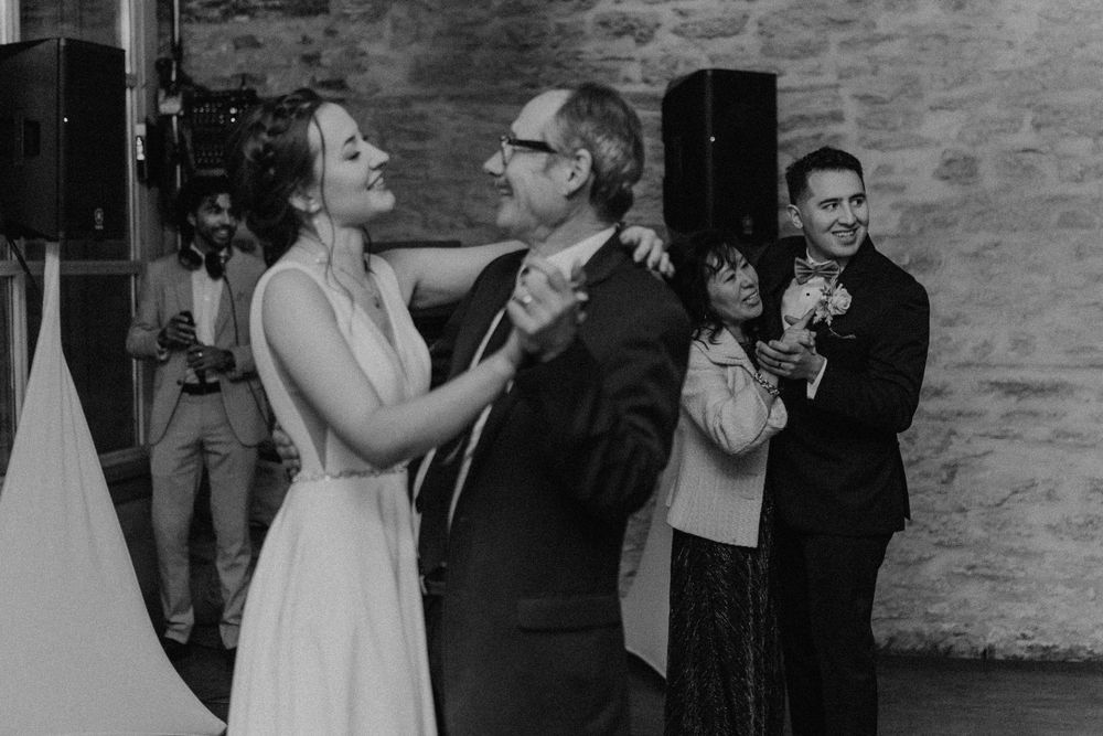 Black and white photo of bride dancing with her father while groom watches joyfully at wedding reception.